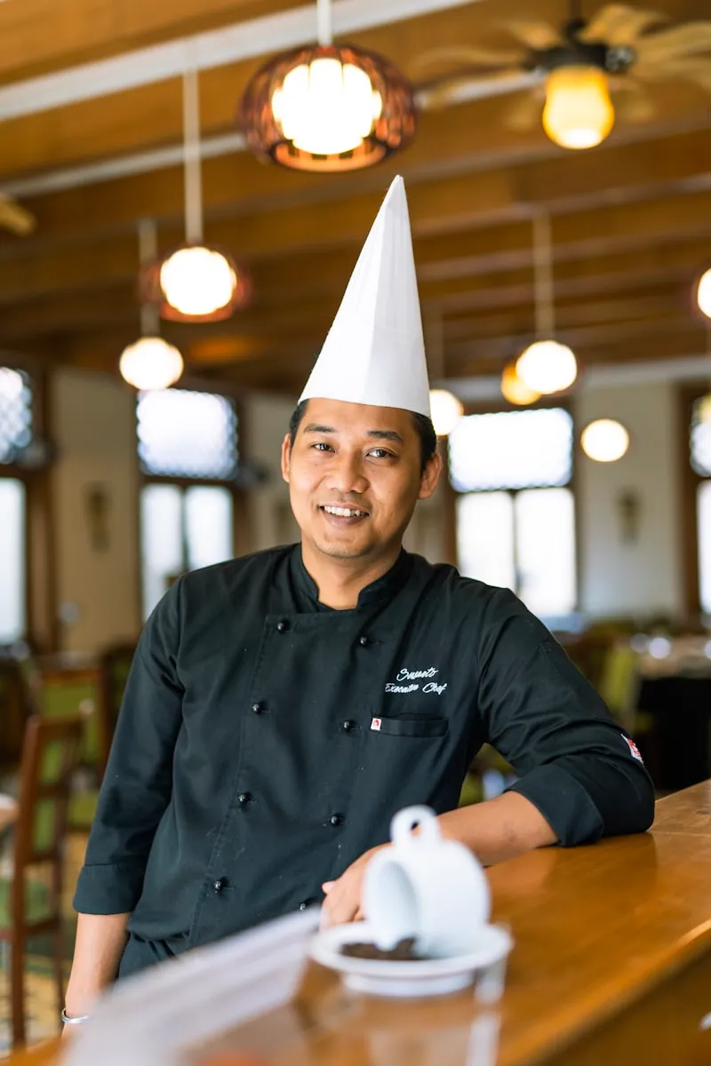 A smiling chef in a toque standing in a restaurant dining room.